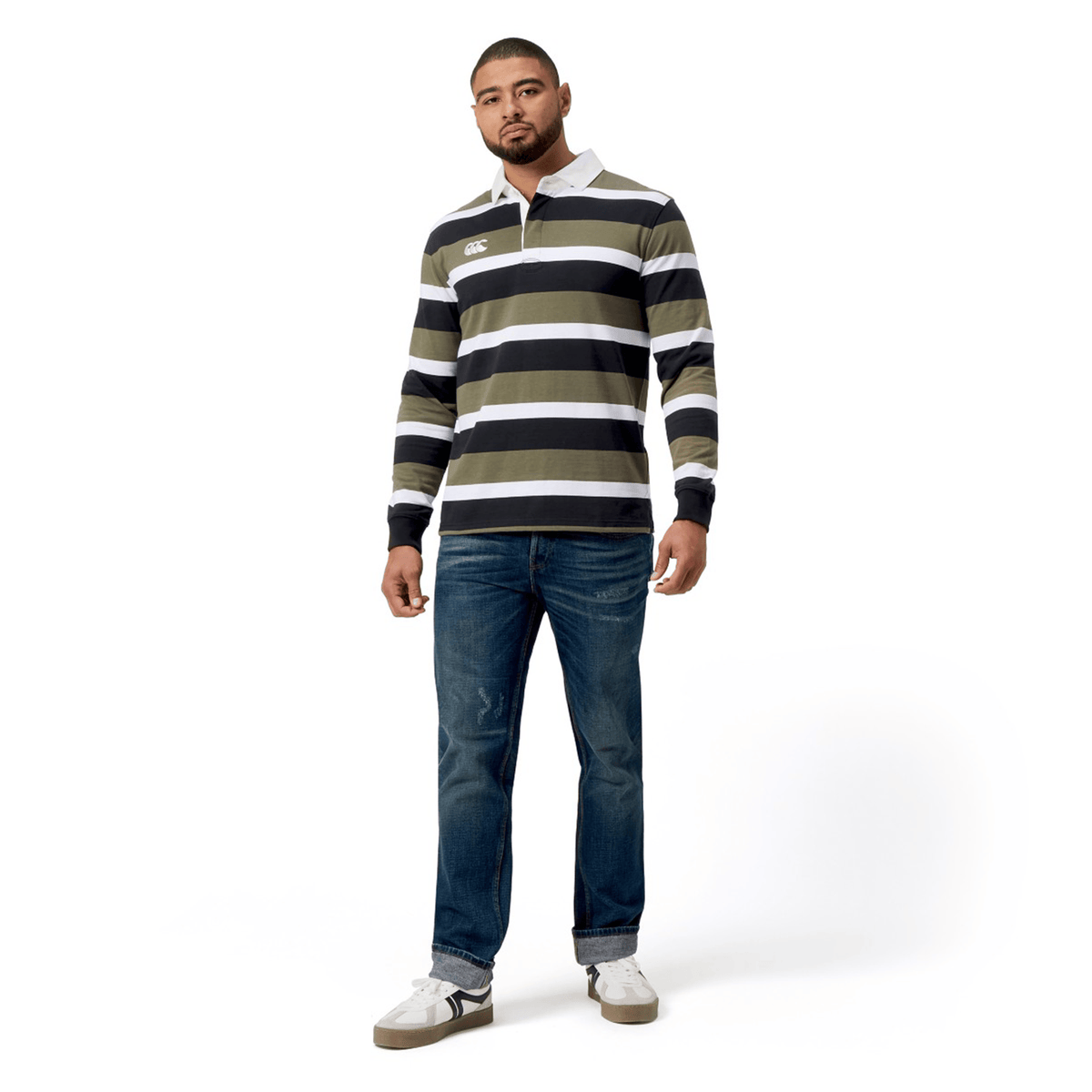 A man stands against a white background wearing the vibrant Canterbury Yarn Dye Long Sleeve Striped Rugby Jersey in green, black, and white, paired with blue jeans and white sneakers.