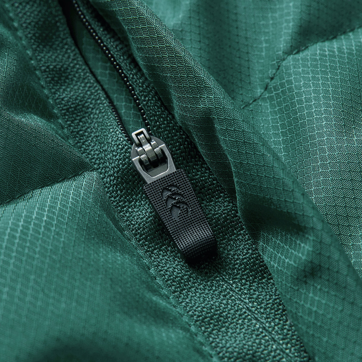 Close-up of a black zipper with a textured pull tab on green ripstop fabric, highlighting detailed stitching and fabric pattern—showcasing the premium craftsmanship of the Ireland 26 Puffer Jacket by Canterbury.
