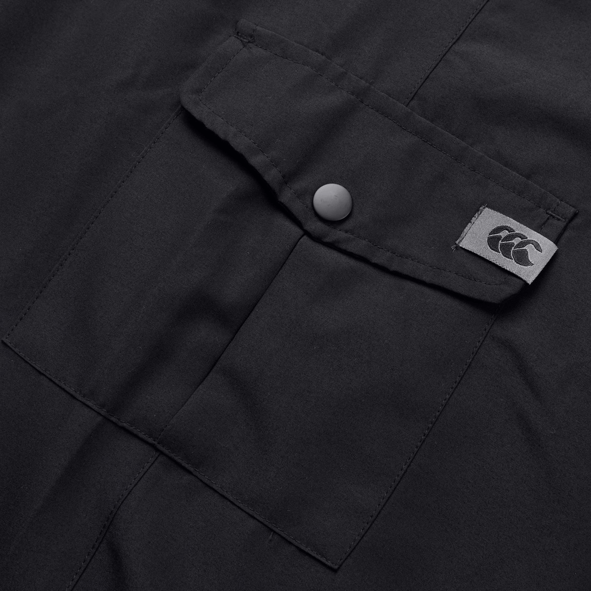 Close-up of black fabric from the Canterbury Force Straight Leg Pant, showcasing a flap pocket with button closure and a small Canterbury logo tag.
