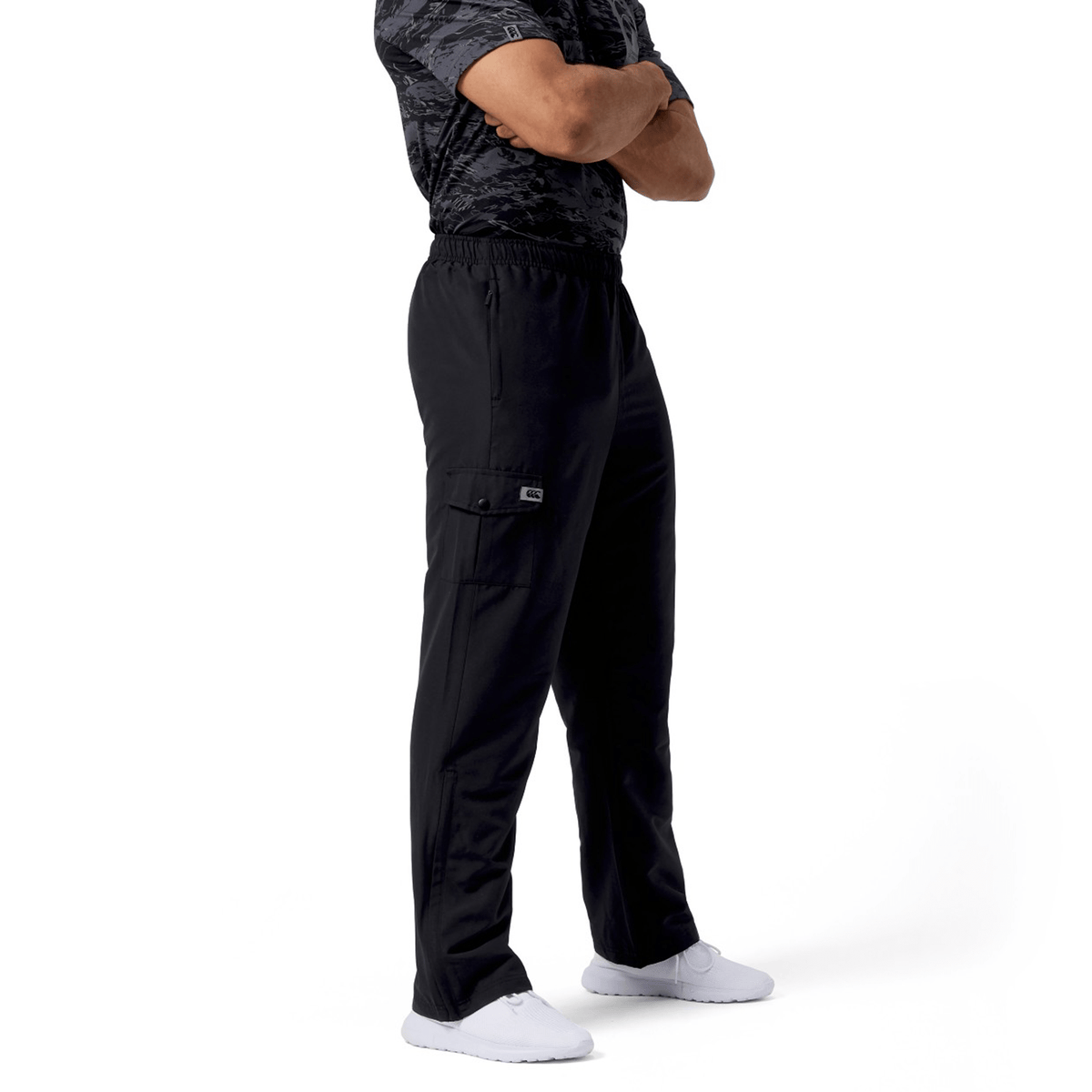 A person stands sideways with arms crossed, wearing a dark camouflage shirt and sleek black Canterbury Force Straight Leg Pant, paired with white sneakers against a white background.