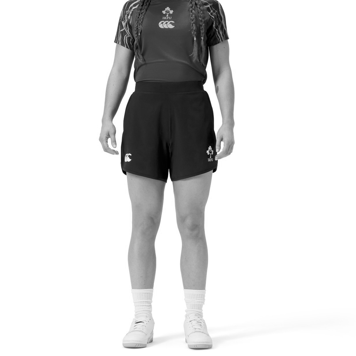 A person wearing a sports uniform stands facing forward, dressed in a short-sleeve shirt, Canterbury Women's Ireland 25/26 Woven Gym Shorts, socks, and sneakers.