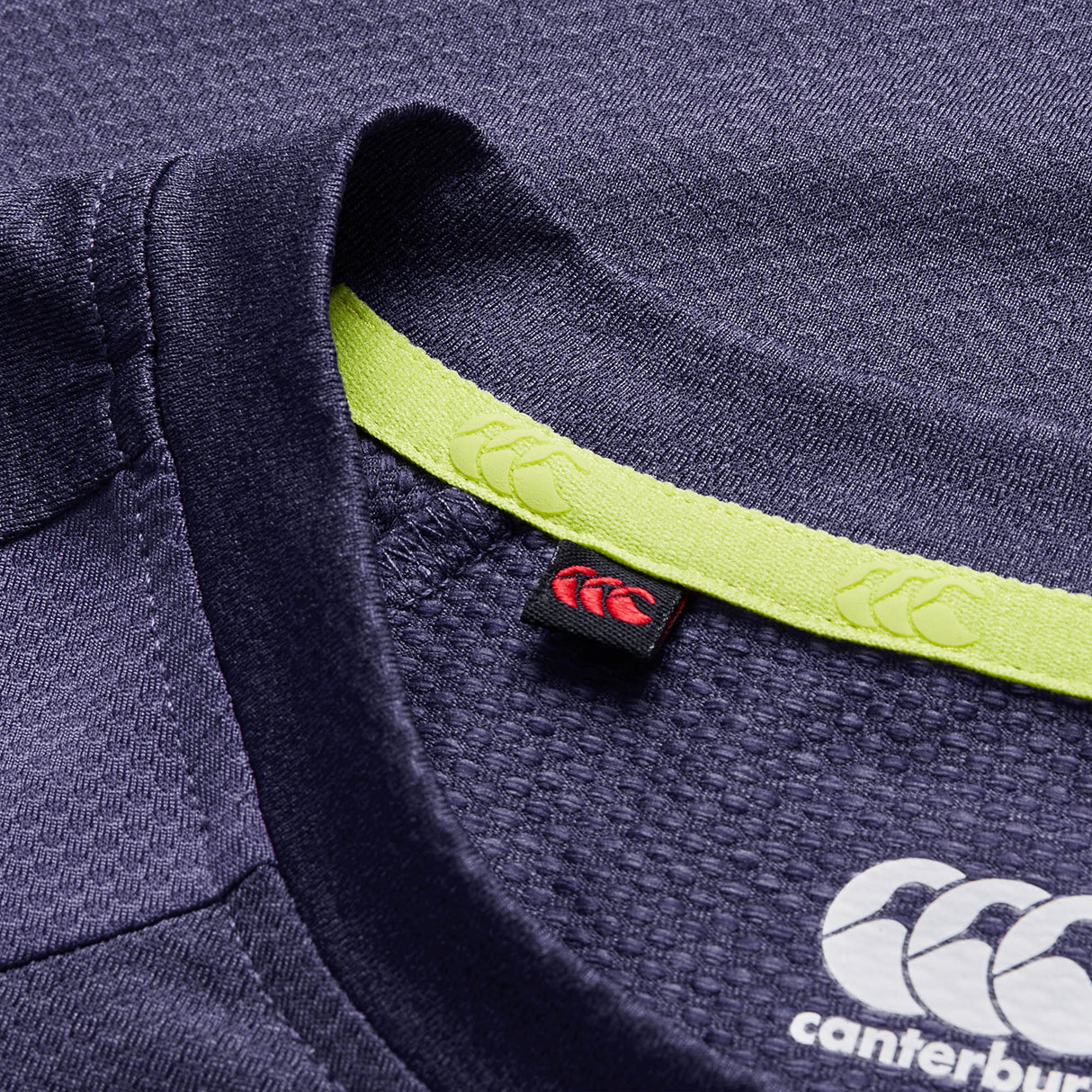 Close-up of the Ireland 26 Elite Training Tee by Canterbury in navy blue, highlighting its yellow inner collar trim, red fabric tag, and white printed Canterbury logo. This Vapodri breathable sportswear top is performance-designed, reflecting the style of official Ireland rugby training apparel.