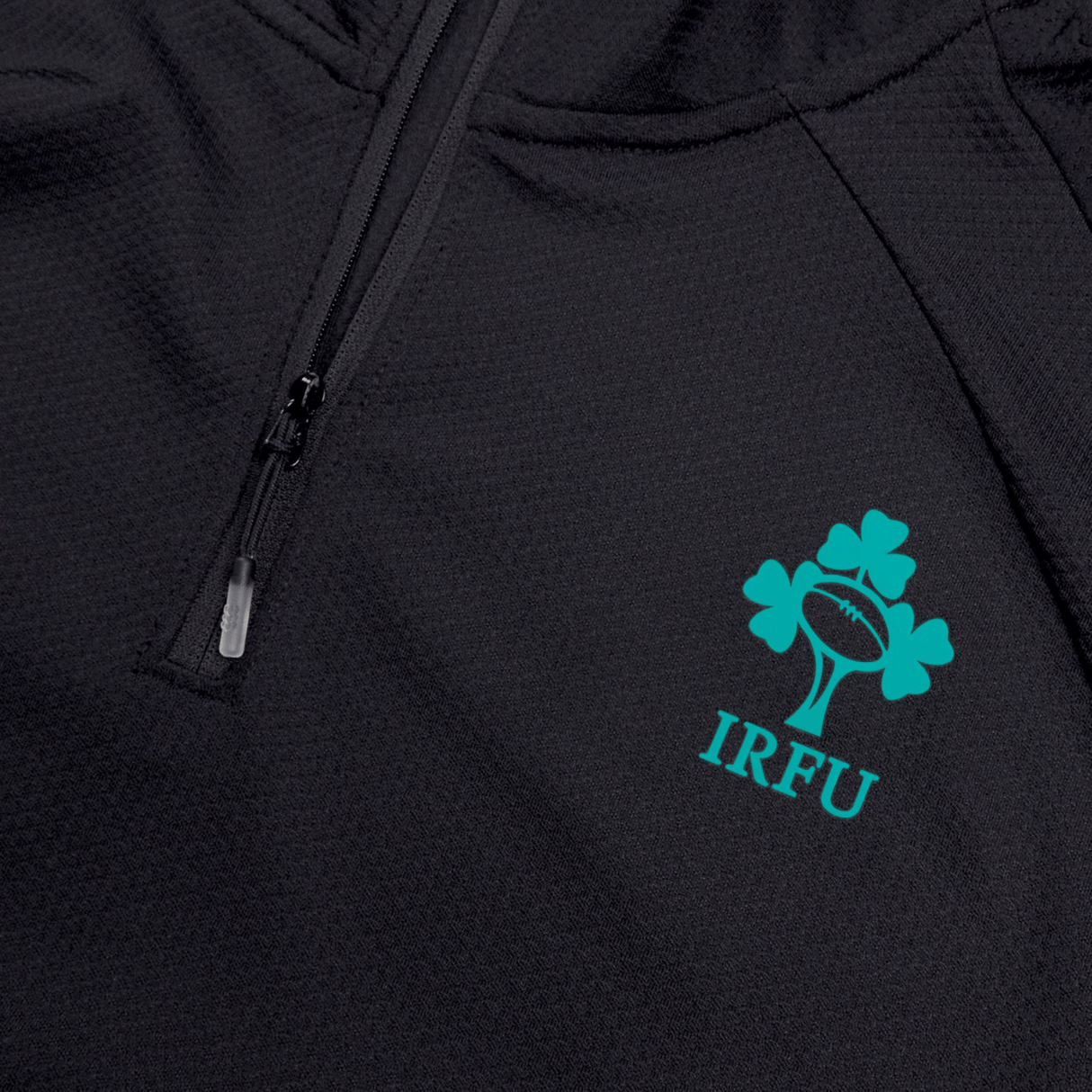 Close-up of the Ireland 25 Elite First Layer by Canterbury, a black zip-up training top featuring the IRFU shamrock and rugby ball logo in teal.