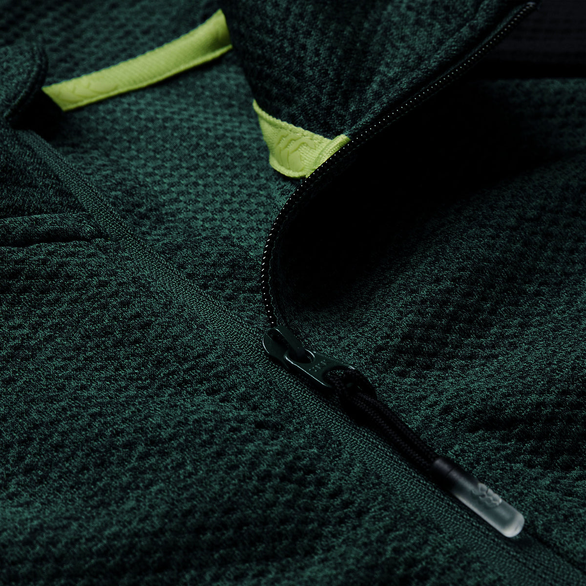 Close-up of the Ireland 26 Elite First Layer by Canterbury, a textured dark green fleece jacket inspired by an Ireland Rugby training top, featuring a partially opened black zipper and a yellow fabric accent near the collar.