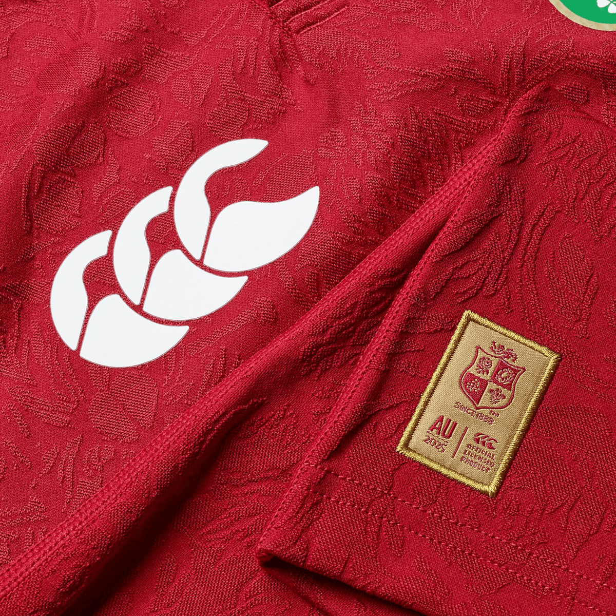 Close-up of a red British & Irish Lions 2025 Youth Players Jersey by Canterbury, crafted from moisture-wicking fabric, showcasing a white logo on the left and a rectangular patch with a shield and text on the right.