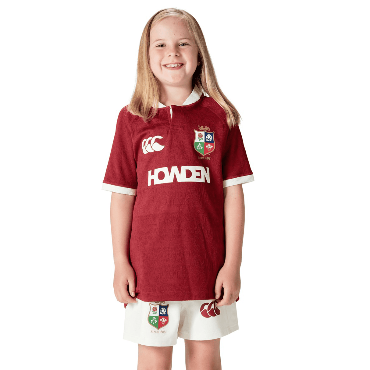 A young girl with long blonde hair smiles, wearing a Youth British & Irish Lions 2025 Players Jersey by Canterbury. The maroon and white rugby jersey and shorts feature logos and moisture-wicking fabric, perfect for active days.