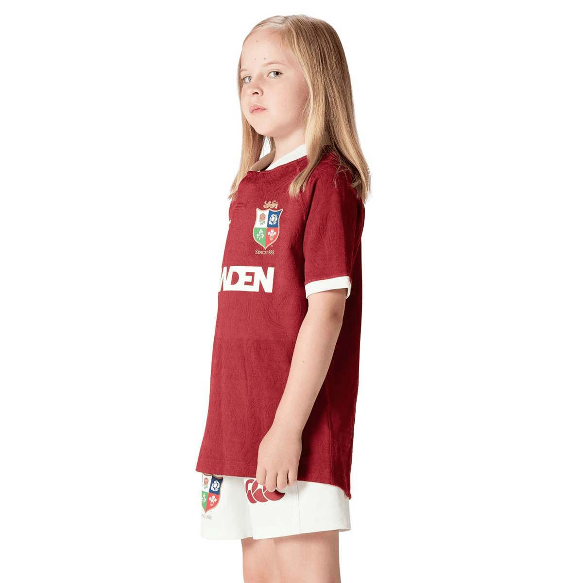 A young girl confidently stands wearing the Youth British & Irish Lions 2025 Players Jersey by Canterbury, featuring moisture-wicking fabric and ergonomic paneling for style and comfort, as she gazes thoughtfully to the side.