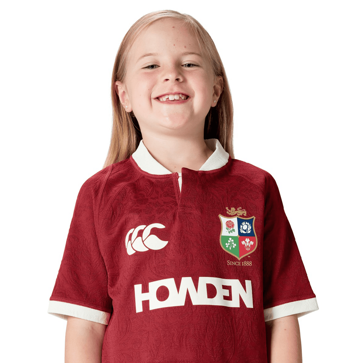 A smiling child proudly wears the maroon Youth British & Irish Lions 2025 Players Jersey by Canterbury, which offers ergonomic paneling for comfort and a moisture-wicking fabric to keep them dry during active play.