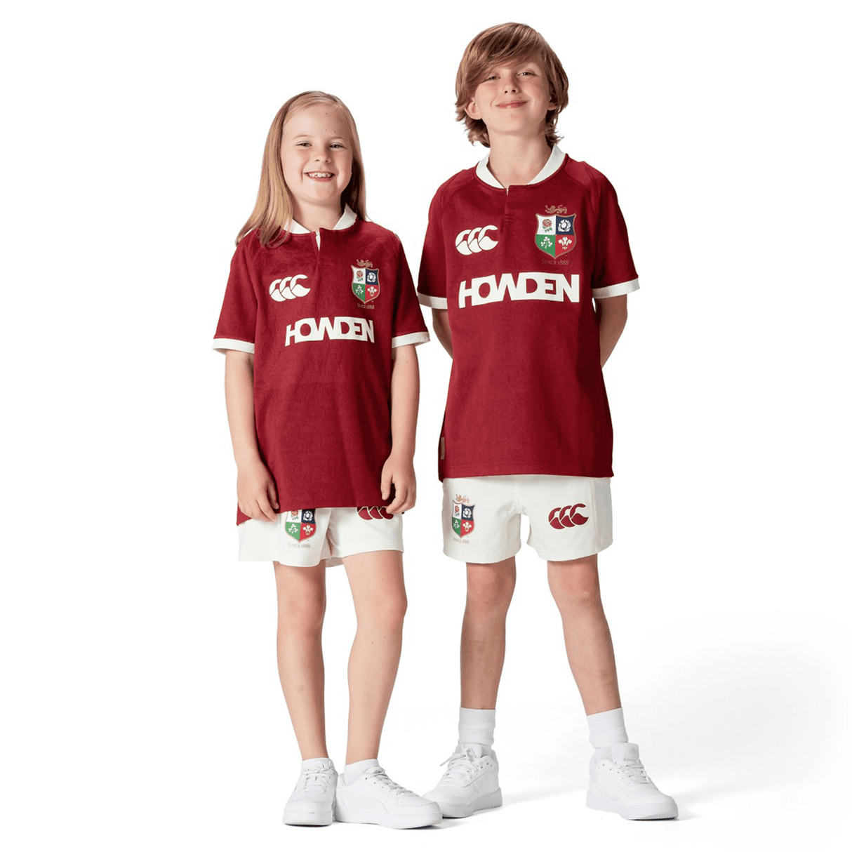 Two smiling children wearing matching red Youth British & Irish Lions 2025 Players Jerseys by Canterbury and white shorts, stand side by side against a plain white background.