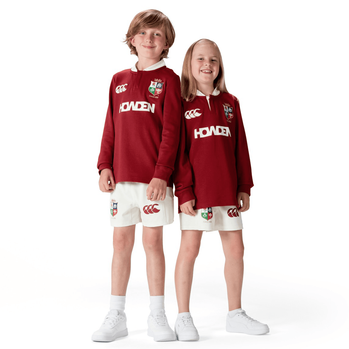 Two children wearing matching Youth British & Irish Lions 2025 Long Sleeve Classic Jerseys by Canterbury in maroon, featuring moisture-wicking fabric and paired with white shorts, stand side by side, smiling.