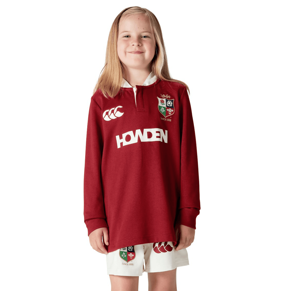 A young girl is smiling and wearing the Youth British & Irish Lions 2025 Long Sleeve Classic Jersey by Canterbury, featuring logos and crafted from moisture-wicking fabric.