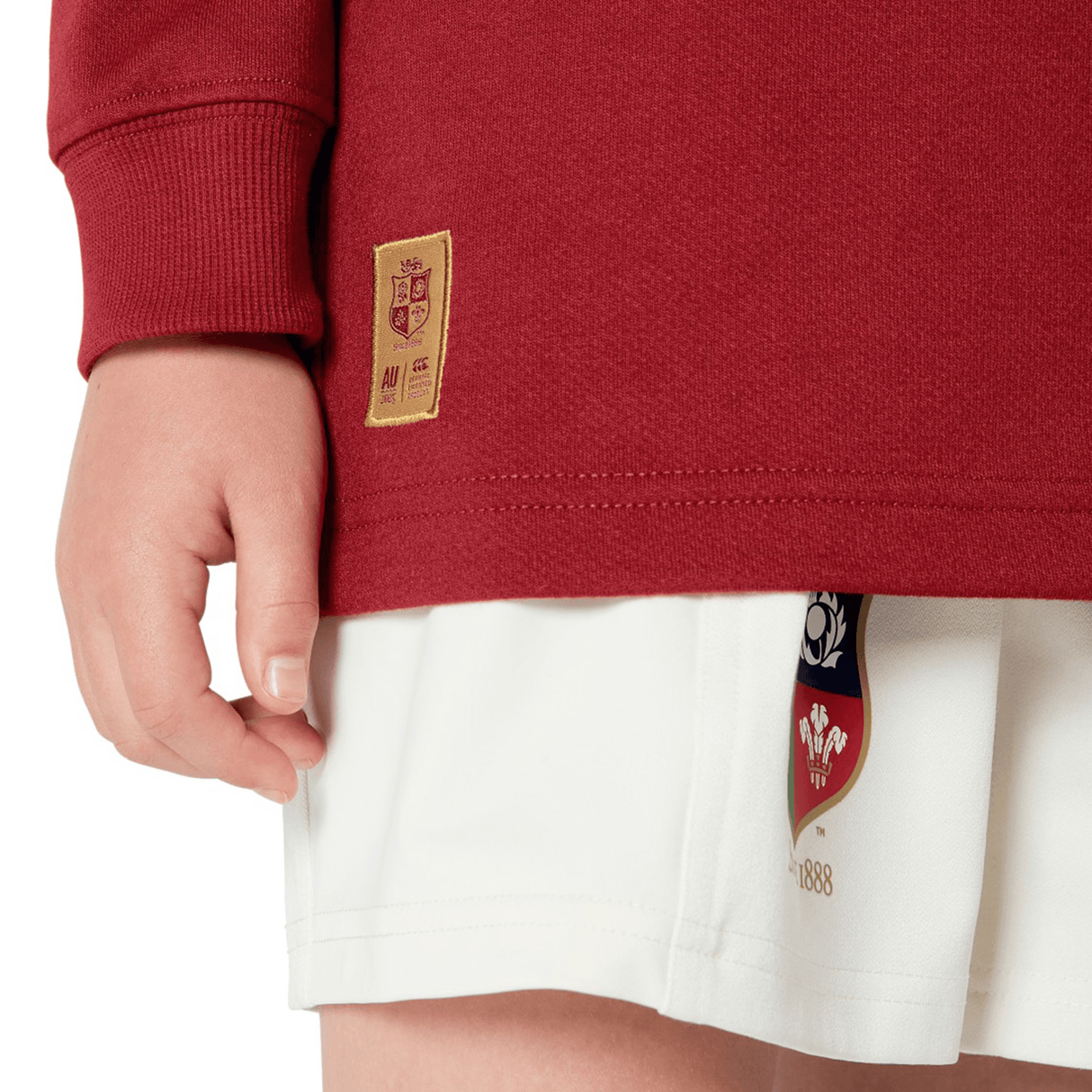 A person models the Youth British & Irish Lions 2025 Long Sleeve Classic Jersey by Canterbury in red with a crest and white shorts, set against a neutral background, highlighting its comfort-driven moisture-wicking fabric.