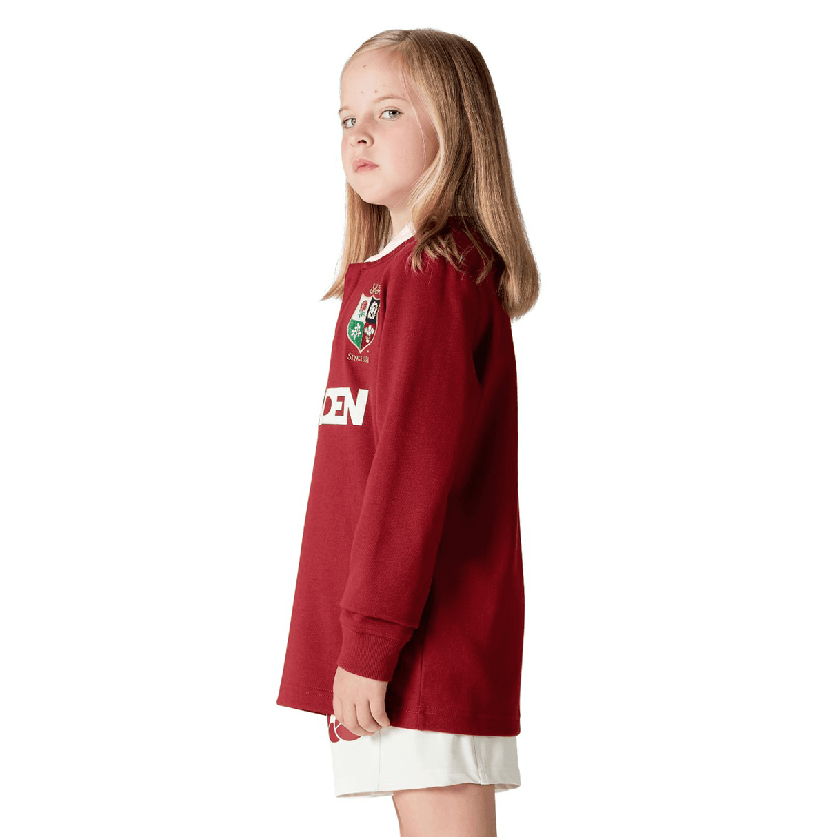 A young girl with long hair stands sideways, wearing the Youth British & Irish Lions 2025 Long Sleeve Classic Jersey by Canterbury and white shorts, looking forward.