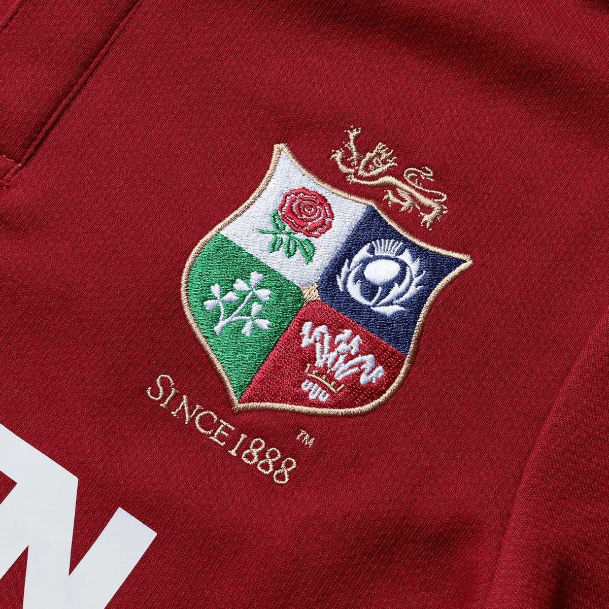 A close-up of the Youth British & Irish Lions 2025 Classic Jersey by Canterbury, featuring a red fabric with an embroidered crest showing a red rose, thistle, shamrock, and dragon. The text "SINCE 1888" honors the legacy of the British & Irish Lions.