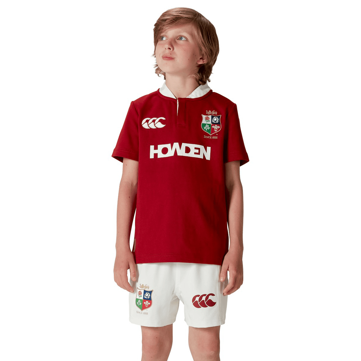 A young boy stands confidently, gazing skyward in a vibrant red Youth British & Irish Lions 2025 Classic Jersey by Canterbury, perfectly paired with logo-adorned white shorts and sleek short sleeves.