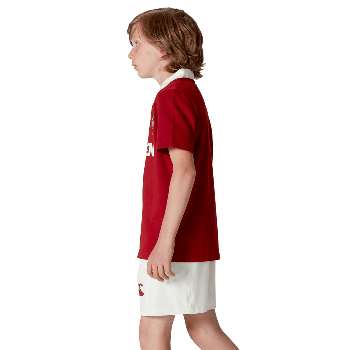 A child with brown hair stands in profile wearing the Youth British & Irish Lions 2025 Classic Jersey by Canterbury in red, paired with white shorts, set against a plain white backdrop.