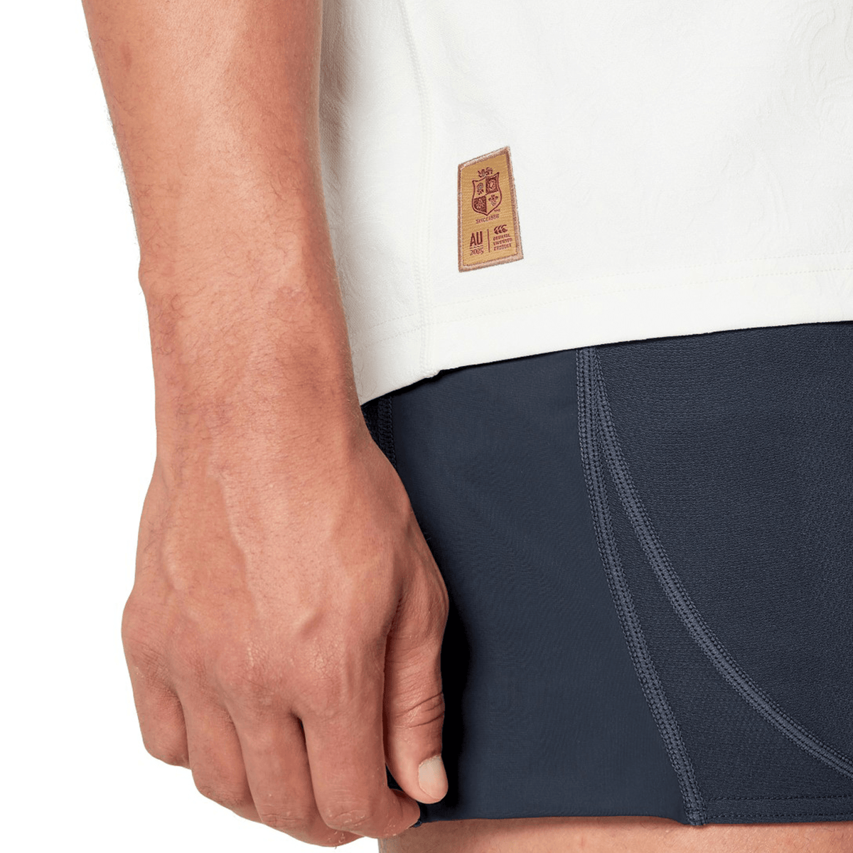 Close-up of a person’s hand resting by their side, wearing a white top with Canterbury branding and dark athletic shorts featuring visible stitching. A small label on the top is reminiscent of the British & Irish Lions 2025 Training Jersey by Canterbury, designed with ergonomically placed panels for comfort.