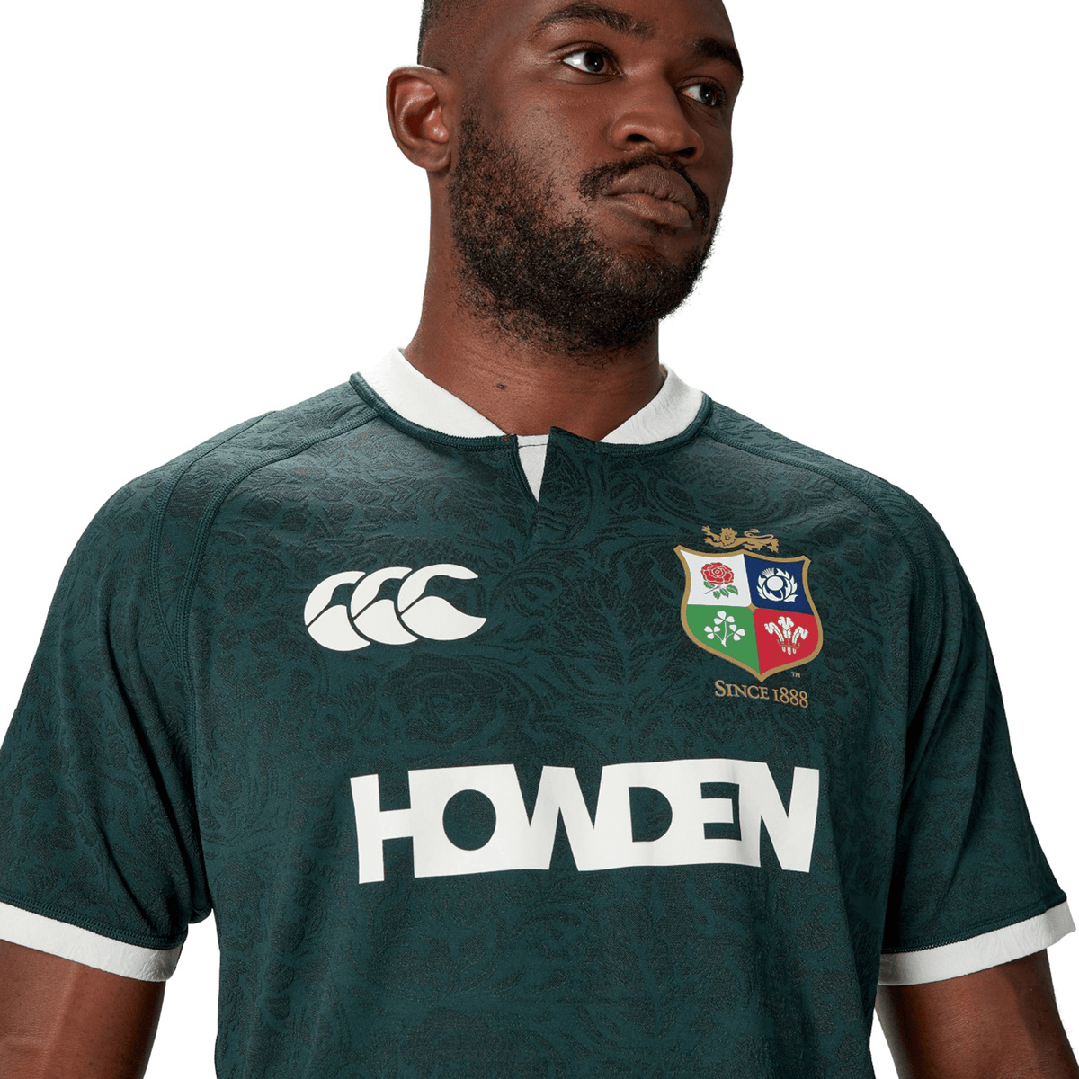A man wearing the British & Irish Lions 2025 Training Jersey by Canterbury, featuring logos and "Howden" text. This stylish, dark green piece is crafted from recycled polyester for an eco-friendly touch.