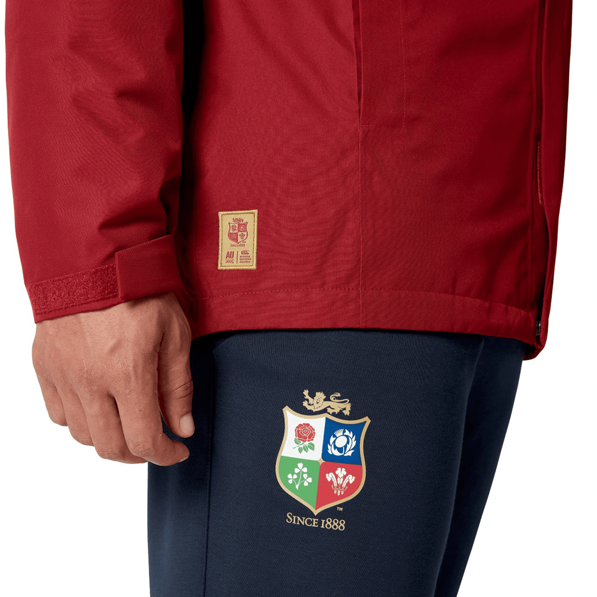 A person dressed in a British & Irish Lions 2025 Waterproof Jacket by Canterbury, featuring an adjustable hood in red, along with dark pants adorned with a multi-colored crest logo and the phrase "Since 1888.