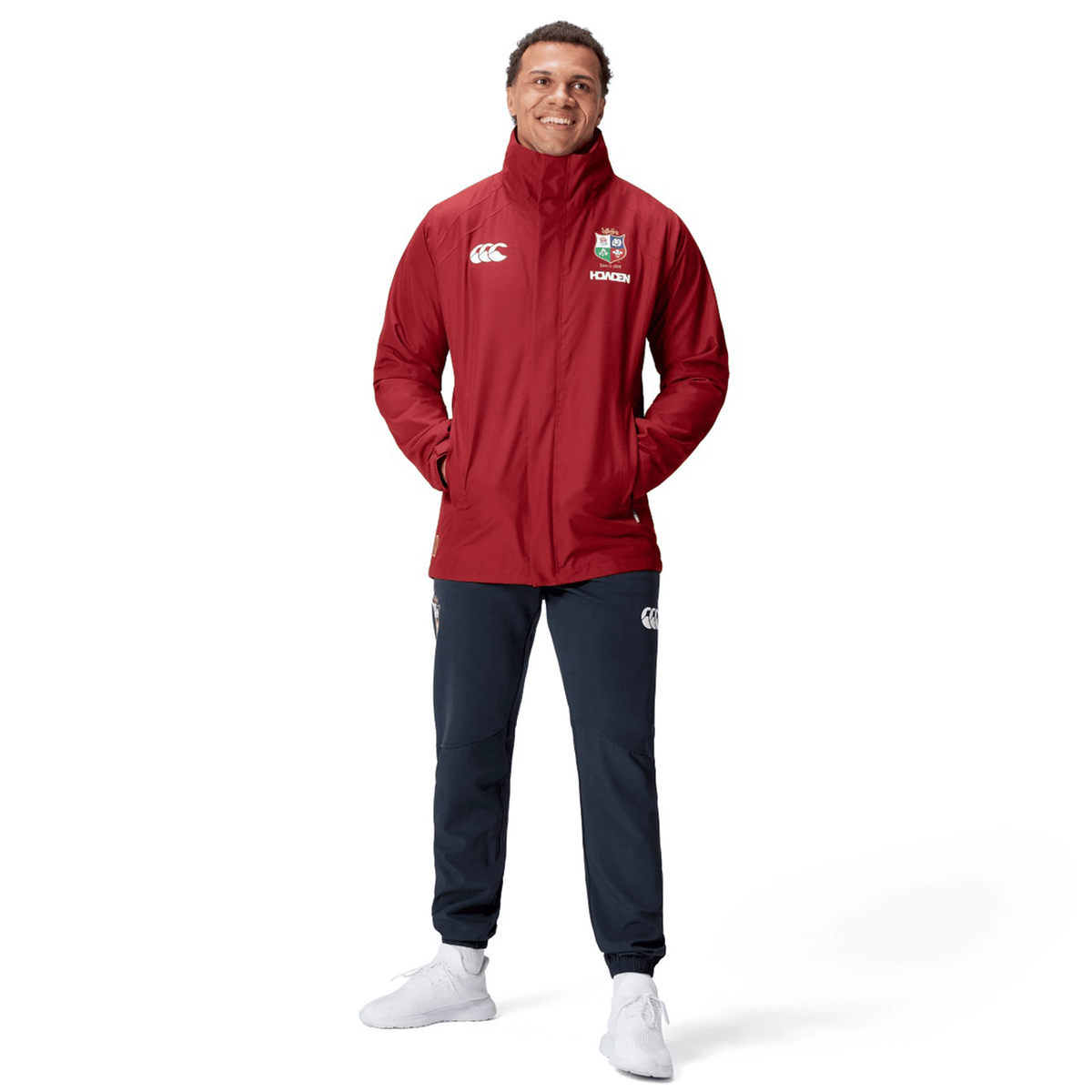 A person in a British & Irish Lions 2025 Waterproof Jacket by Canterbury, featuring logos, stands smiling with hands in pockets, wearing black pants and white sneakers against a white background.