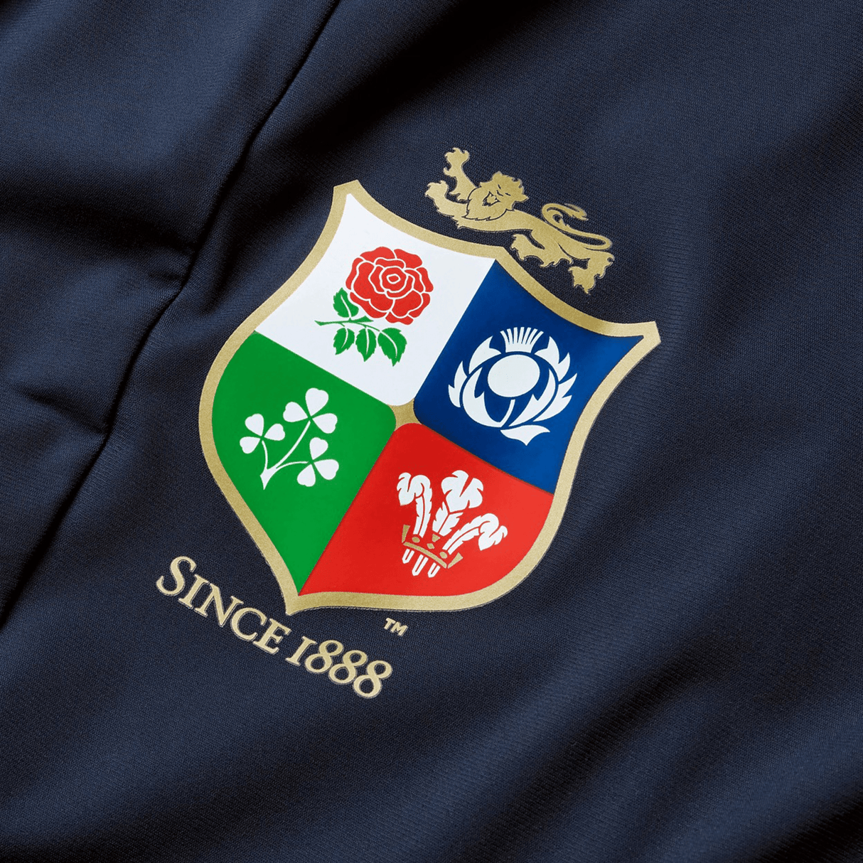The British & Irish Lions 2025 Everest Woven Pant by Canterbury is designed with a navy fabric displaying a coat of arms that includes a lion, rose, thistle, shamrock, and three feathers. The text "Since 1888" highlights the heritage of the British & Irish Lions. Made from quick-drying fabric, it offers both durability and comfort.