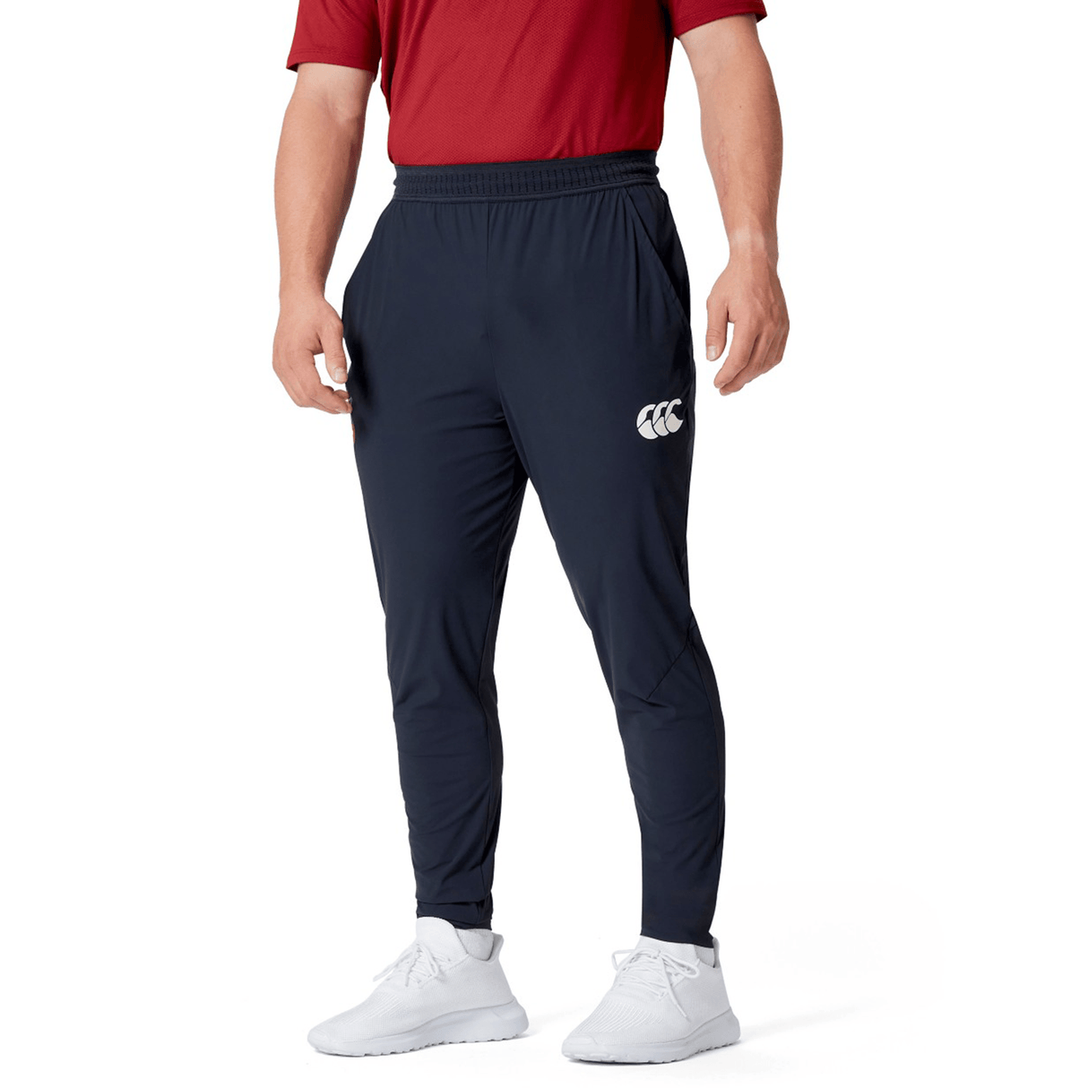 The person sports a red shirt paired with the British & Irish Lions 2025 Everest Woven Pant by Canterbury, which features four-way stretch for optimum comfort. White sneakers complete the look, adding a touch of style and practicality. With hands casually tucked in pockets, they're set for quick-drying adventures.