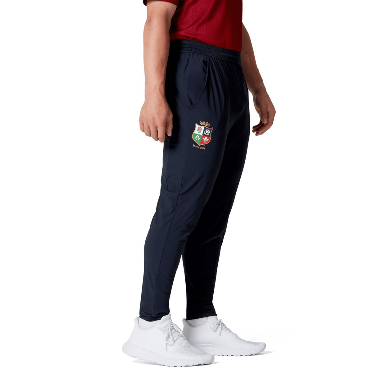 A person in a red shirt wearing the British & Irish Lions 2025 Everest Woven Pant by Canterbury, featuring a crest emblem and paired with white sneakers, stands sideways. This outfit's four-way stretch fabric delivers comfort and quick-drying convenience, ideal for maintaining an active lifestyle.