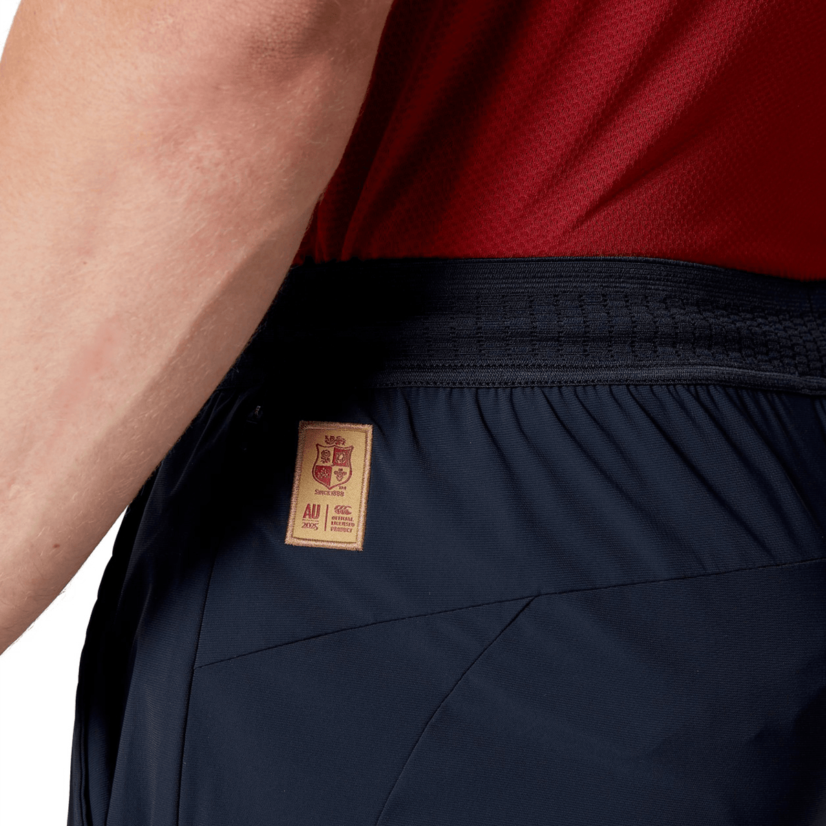 Close-up of a person donning a red shirt and the British & Irish Lions 2025 Everest Woven Pant by Canterbury in navy, designed with quick-drying fabric and featuring a brown label on the waistband, perfect for channeling your inner British & Irish Lions spirit.
