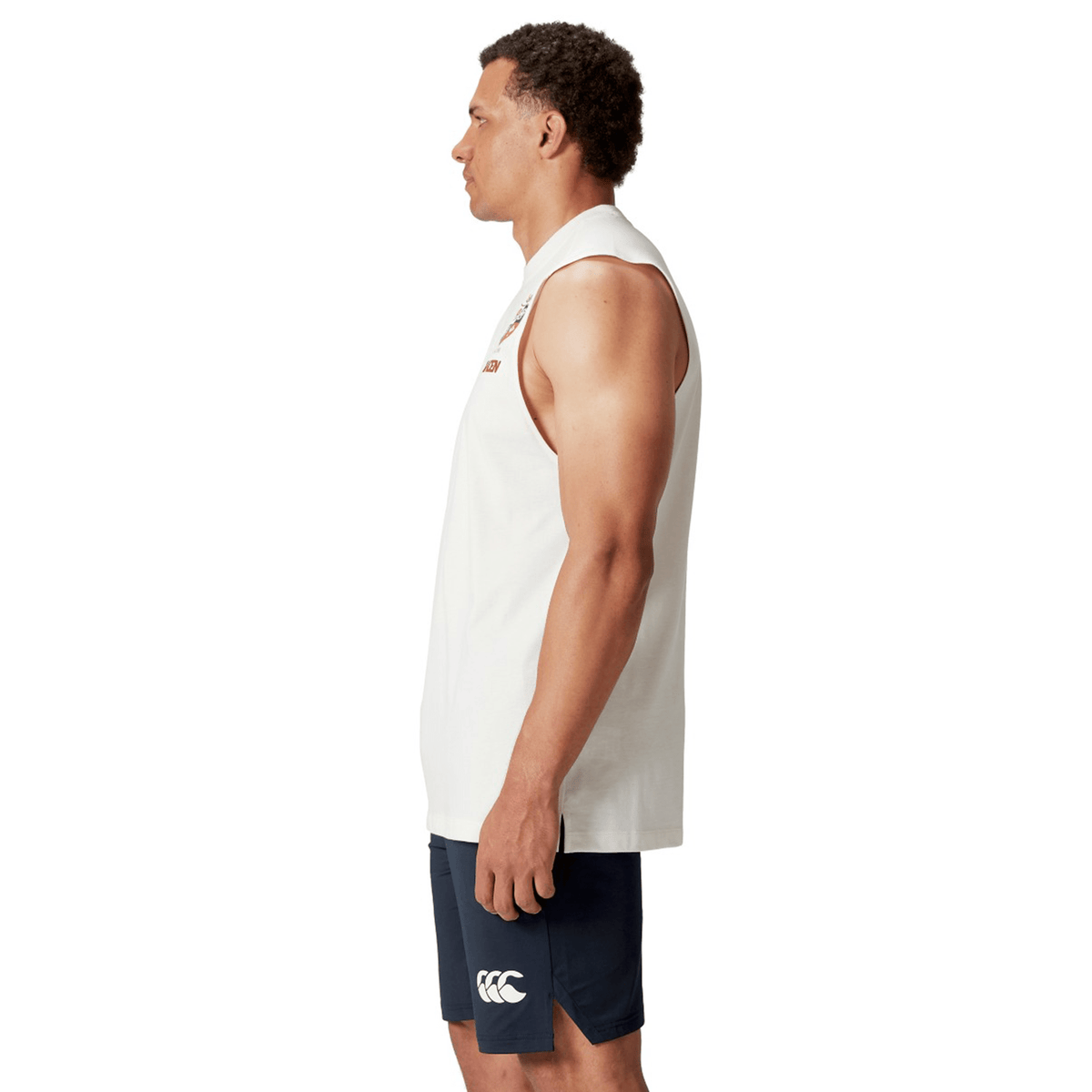 A man stands in profile wearing a British & Irish Lions 25 Cotton Muscle Tank by Canterbury, featuring logos, along with navy shorts, set against a plain background.