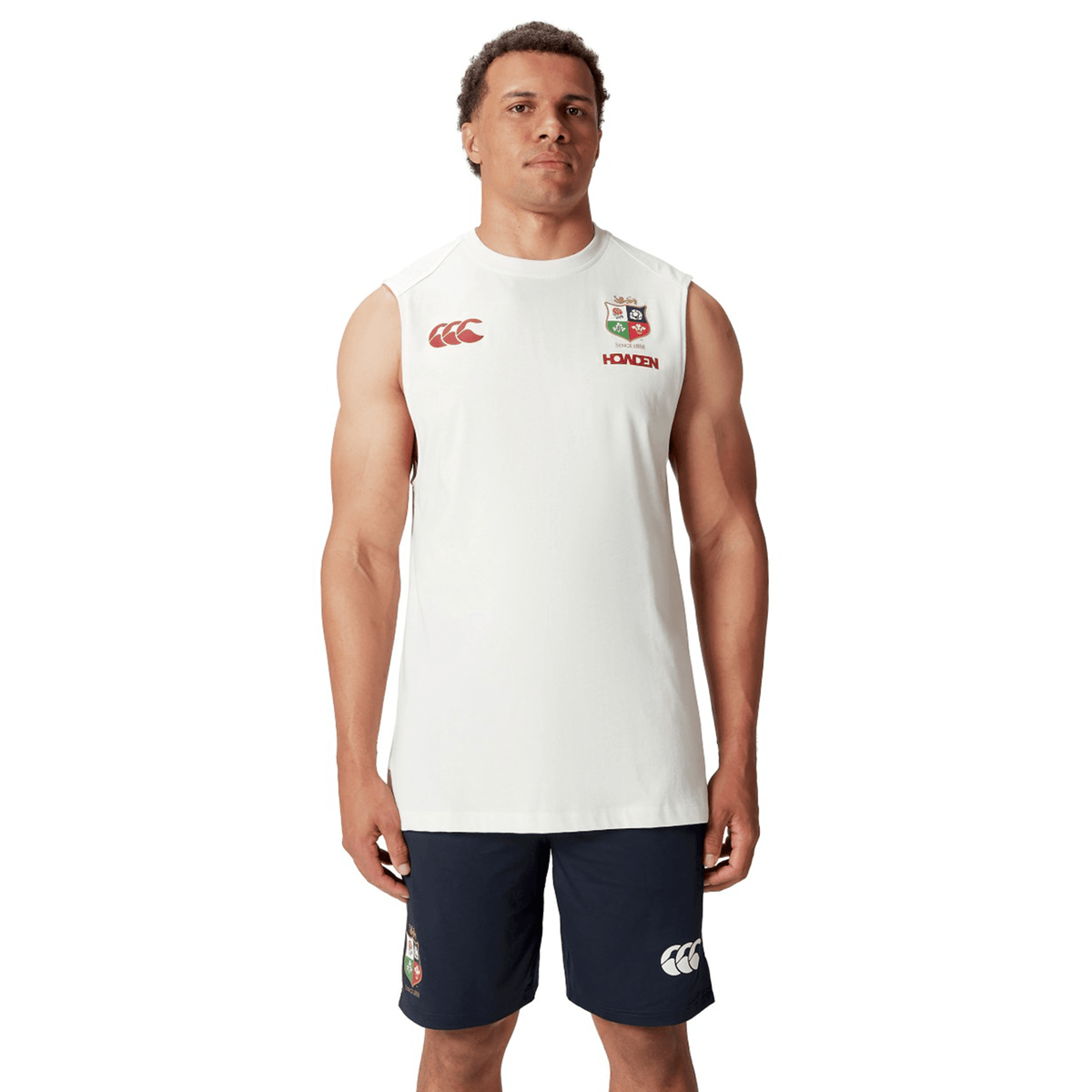 A person wearing a white British & Irish Lions 25 Cotton Muscle Tank by Canterbury and navy shorts with sports logos stands against a white background.
