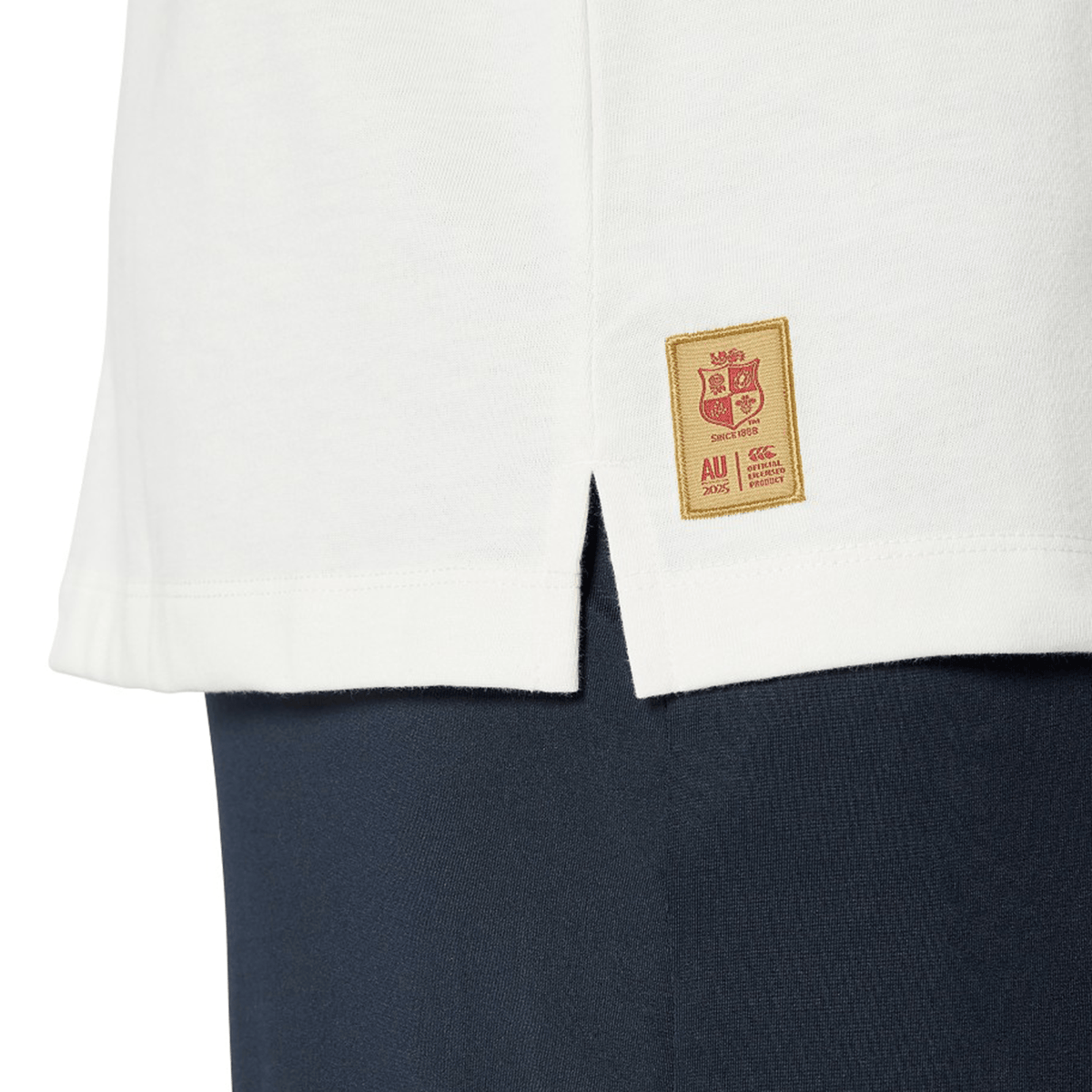 Close-up of the hem of a Canterbury British & Irish Lions 25 Cotton Muscle Tank in white, featuring a small emblem patch above the side slit, worn over dark pants.