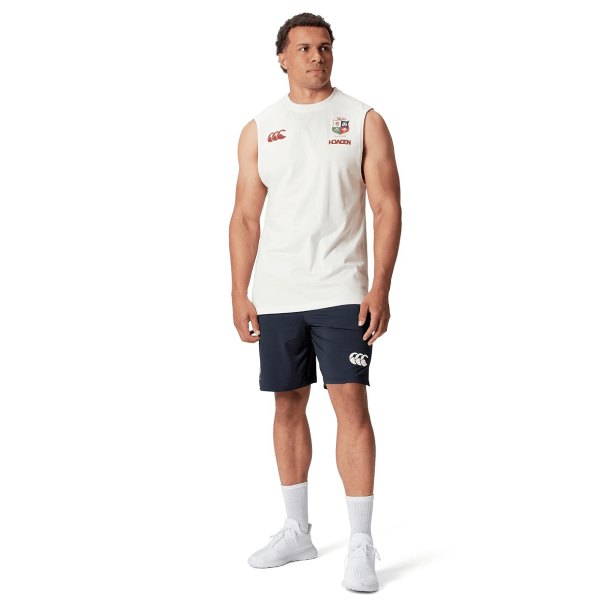 A man stands confidently against a white backdrop, exuding the spirit of the British & Irish Lions, wearing the British & Irish Lions 25 Cotton Muscle Tank by Canterbury and navy shorts.
