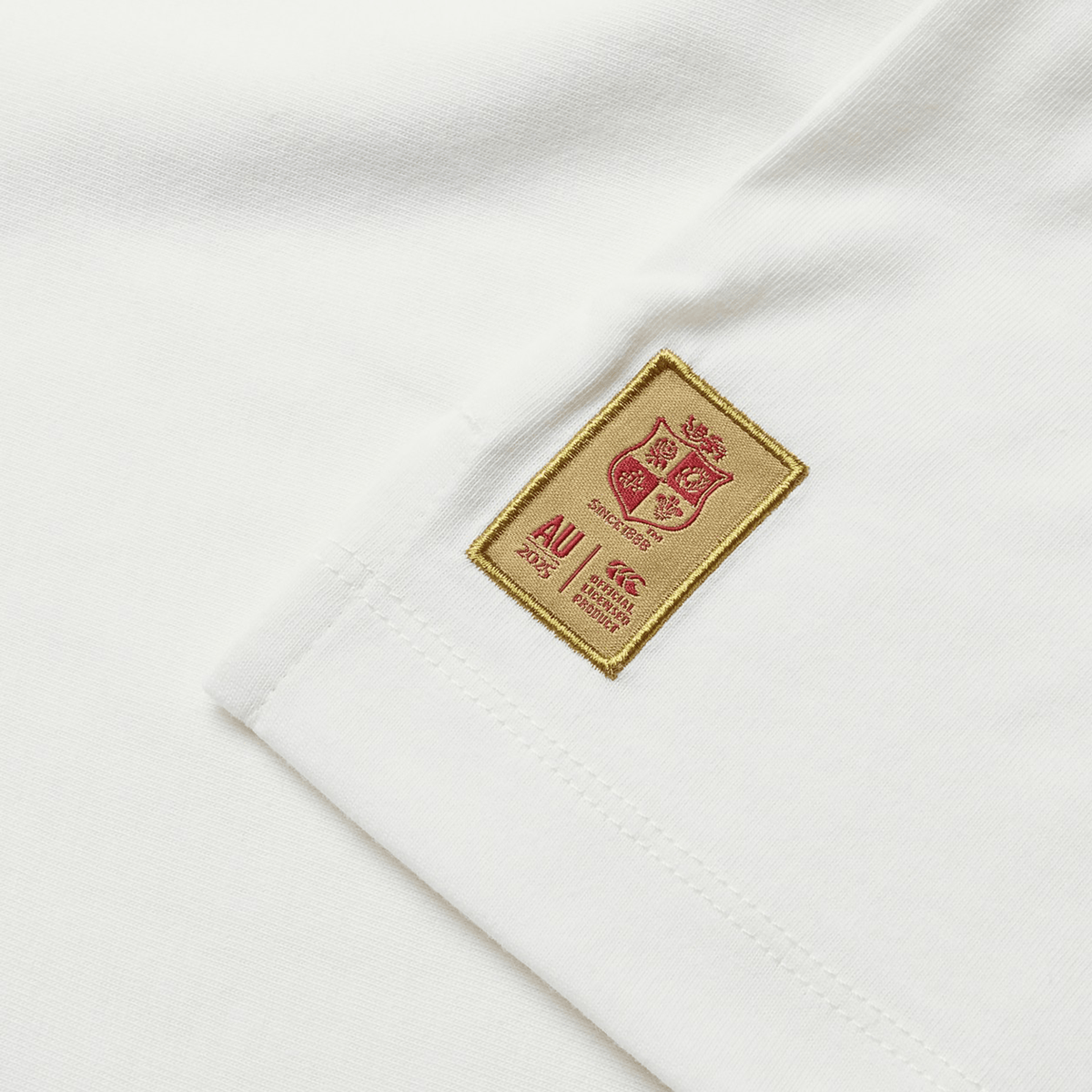 A close-up of a white fabric label on the British & Irish Lions 25 Cotton Muscle Tank by Canterbury displays a red shield emblem and small text. This label, attached to the garment, signifies the esteemed association with Canterbury and the British & Irish Lions.