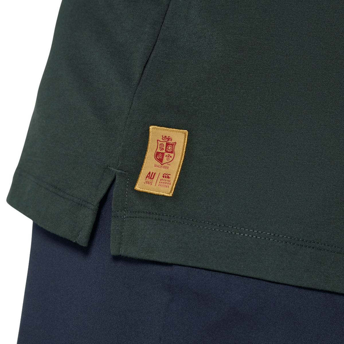 Close-up of the hem of the British & Irish Lions 25 Cotton Muscle Tank by Canterbury in dark green cotton, featuring a small yellow tag with a crest and text, complemented by dark blue pants—a nod to Canterbury's classic style reminiscent of the British & Irish Lions.
