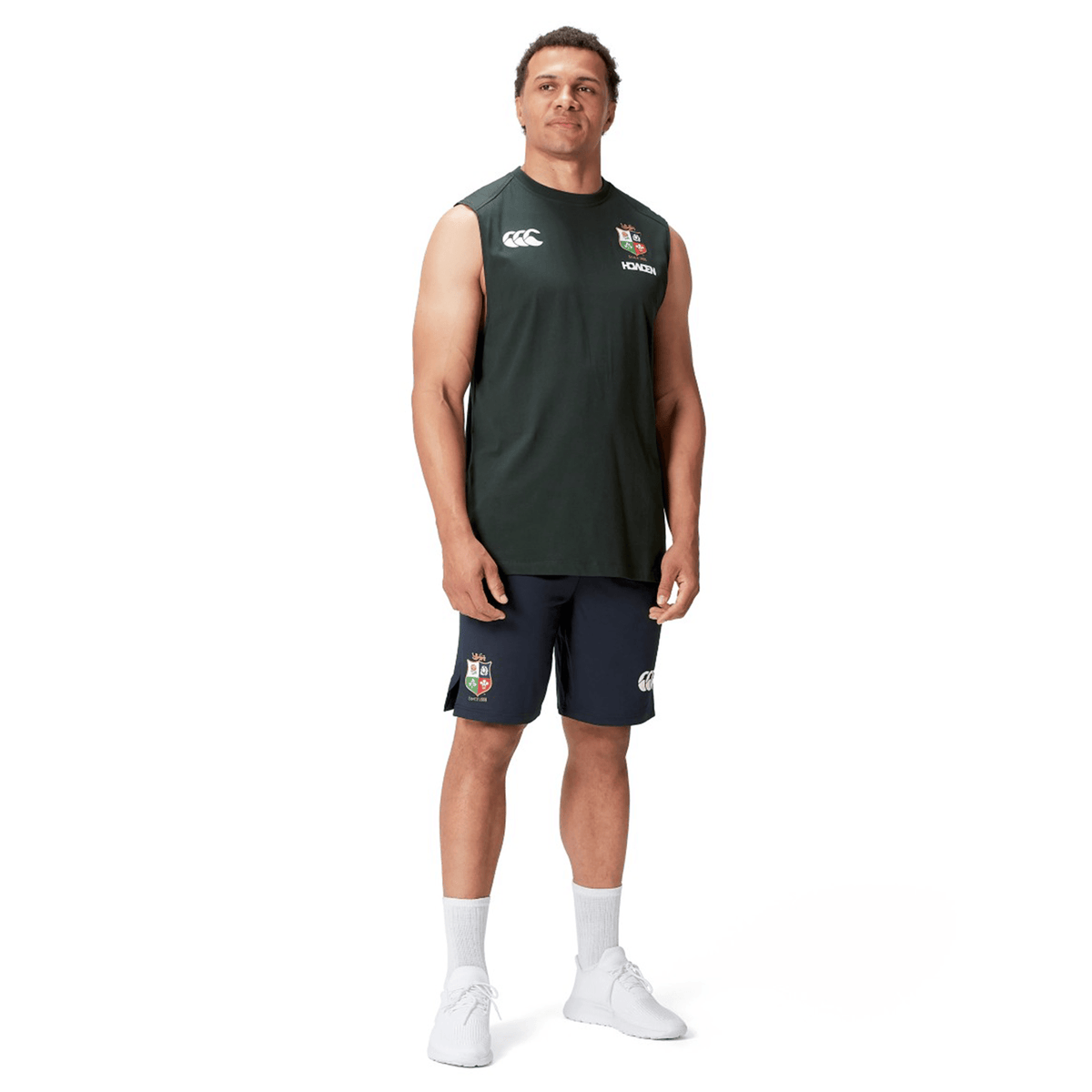A person stands wearing a dark British & Irish Lions 25 Cotton Muscle Tank by Canterbury, navy shorts, white socks, and white sneakers against a plain white background.