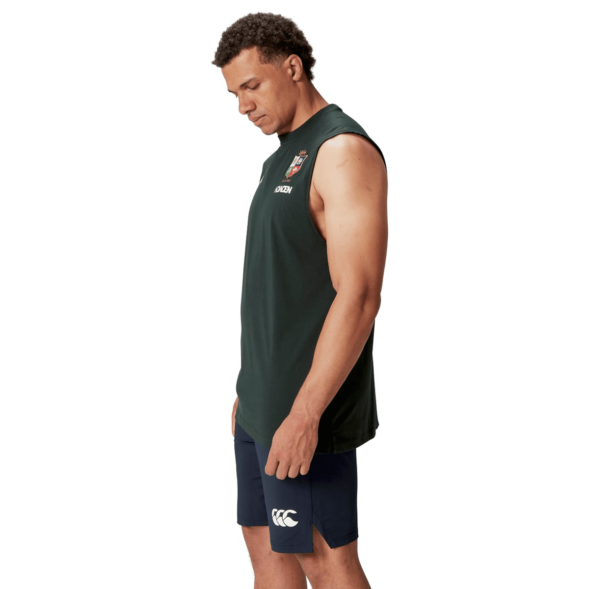 A man wearing a British & Irish Lions 25 Cotton Muscle Tank by Canterbury and navy shorts stands facing right against a white background, capturing the essence of the British & Irish Lions.