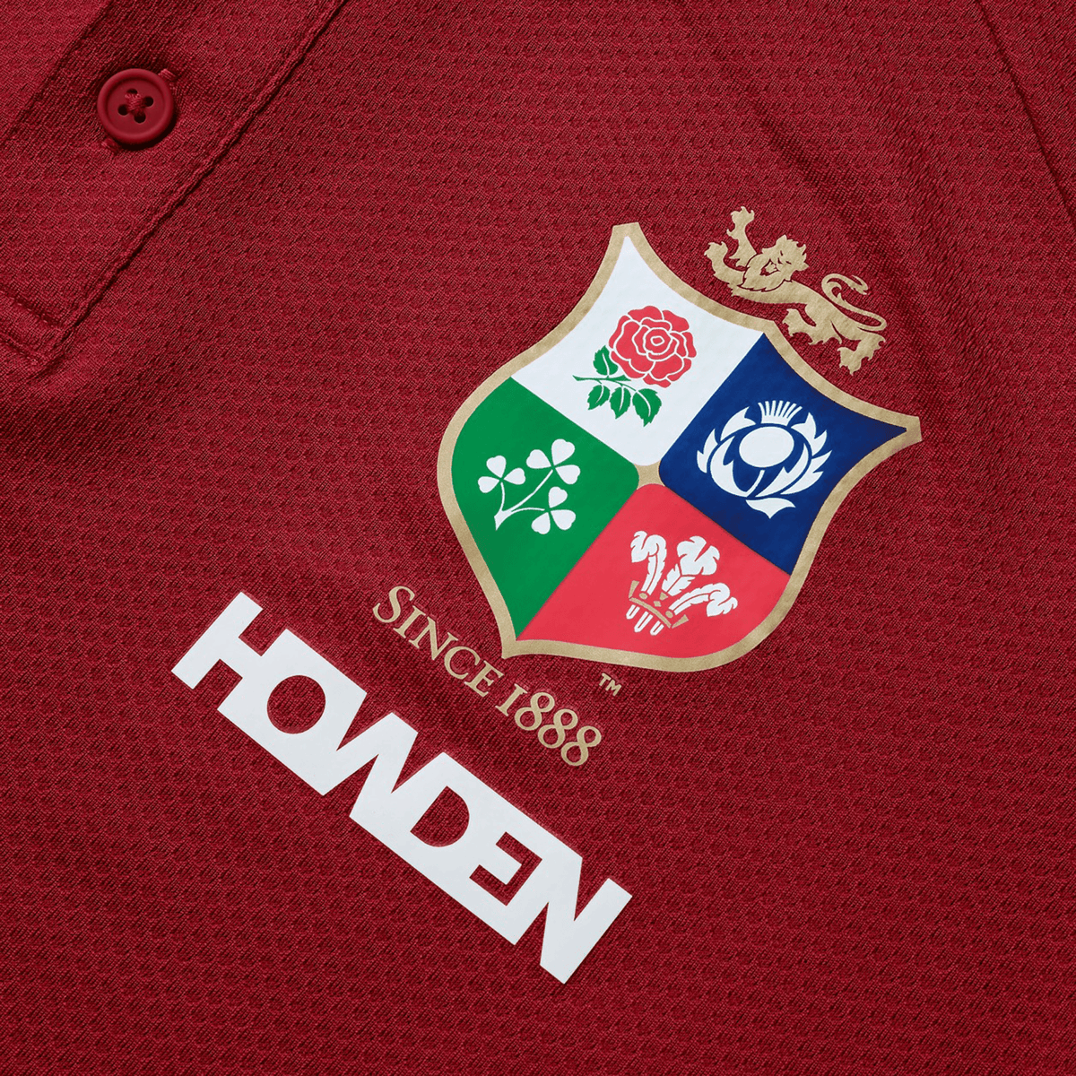 Close-up of a red shirt in breathable wicking fabric, featuring an embroidered shield with a lion, rose, clover, and thistle. Dated "Since 1888," with "HOWDEN" printed below, this design echoes the classic style of the British & Irish Lions 25 Poly Polo by Canterbury.