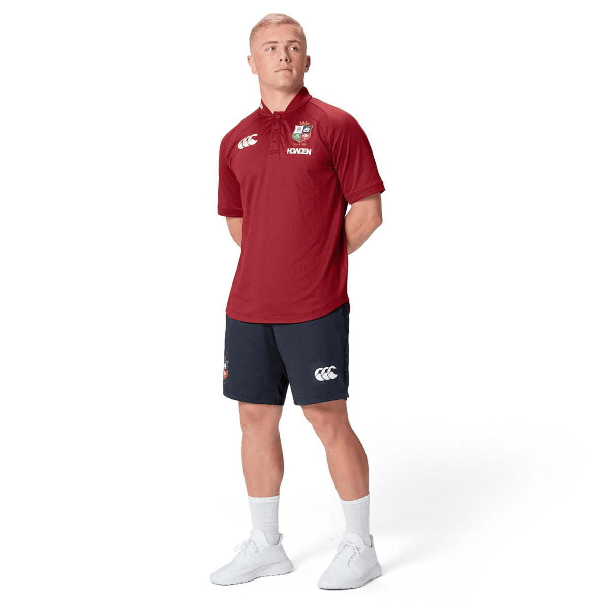 A person wearing the British & Irish Lions 25 Poly Polo by Canterbury, coupled with black shorts, stands with hands behind their back and gazes to the side on a white backdrop, capturing the timeless style of Canterbury and the vibrant heritage of the British & Irish Lions.