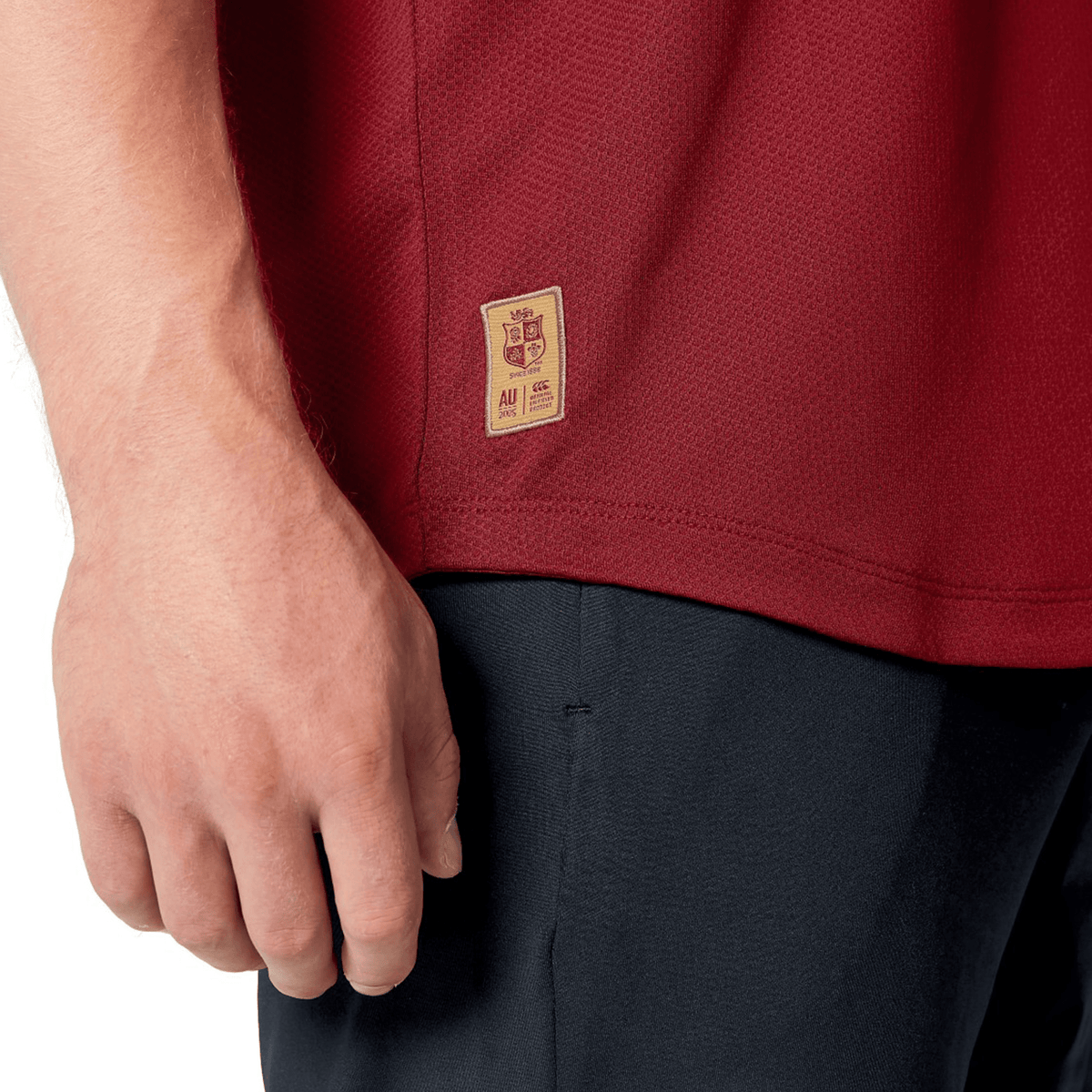 Close-up of a person’s hand by their side, wearing the red British & Irish Lions 25 Poly Polo from Canterbury, featuring breathable fabric and a small tag, paired with black pants.