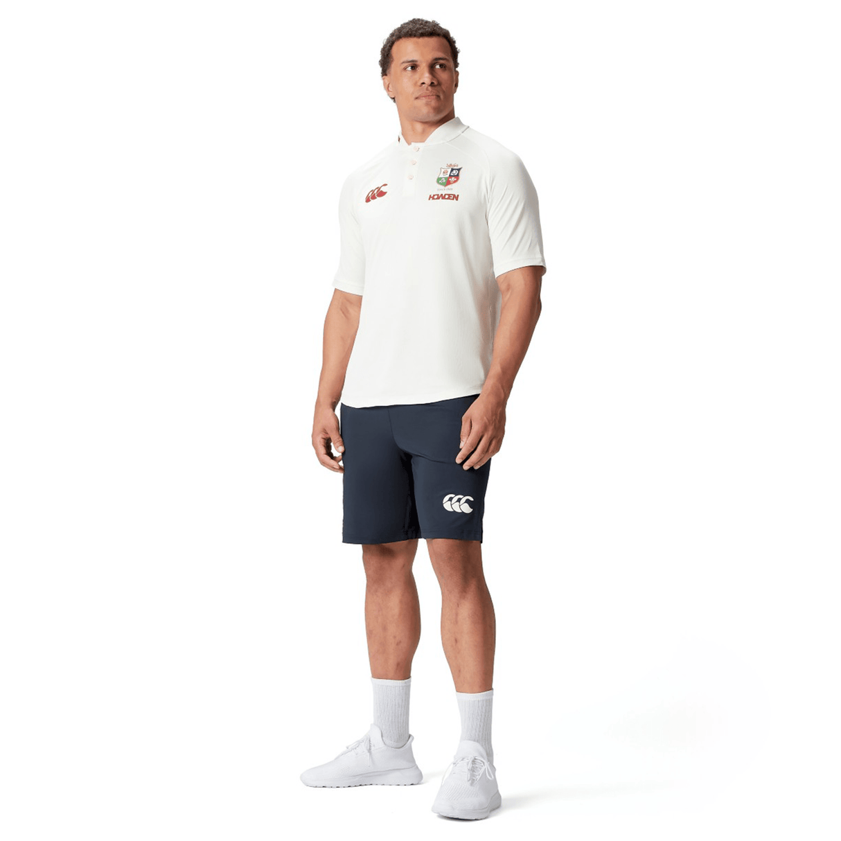 A man in sports attire stands confidently, wearing the British & Irish Lions 25 Poly Polo by Canterbury, paired with navy shorts, white socks, and sleek white sneakers.