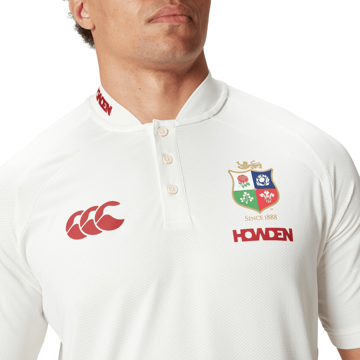 An individual is dressed in the British & Irish Lions 25 Poly Polo by Canterbury, showcasing a white rugby shirt with a red Canterbury logo, the British and Irish Lions crest, and "Howden" branding. Its quick-drying fabric provides comfort during any match.