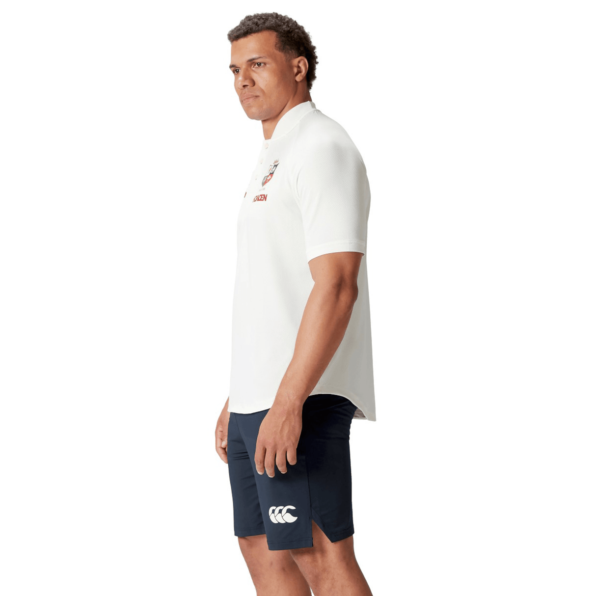 A man wearing a white British & Irish Lions 25 Poly Polo by Canterbury and navy shorts stands against a white background, highlighting the embossed BIL neck tape.