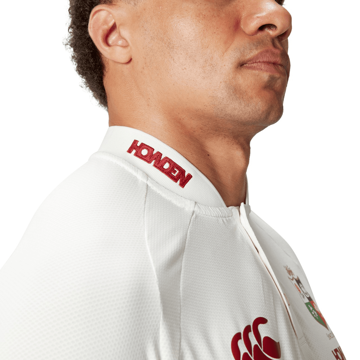 A side profile of a person wearing the British & Irish Lions 25 Poly Polo by Canterbury, a white sports jersey adorned with red logos and text on the collar. Made with quick-drying fabric for enhanced comfort, this design features an embossed BIL neck tape that adds a refined touch.
