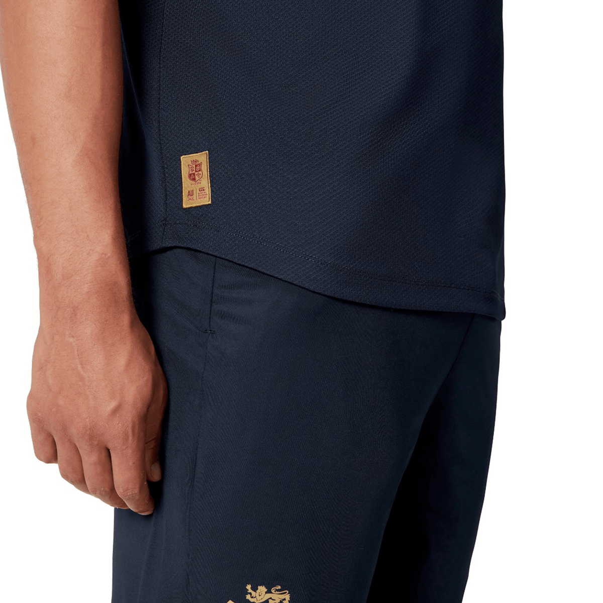 A close-up shot shows someone wearing the British & Irish Lions 25 Poly Polo by Canterbury in a dark blue color with gold details. The quick-drying fabric of the polo is highlighted, along with their left hand positioned near the side seam logo on the shirt, showcasing Canterbury's commitment to quality.