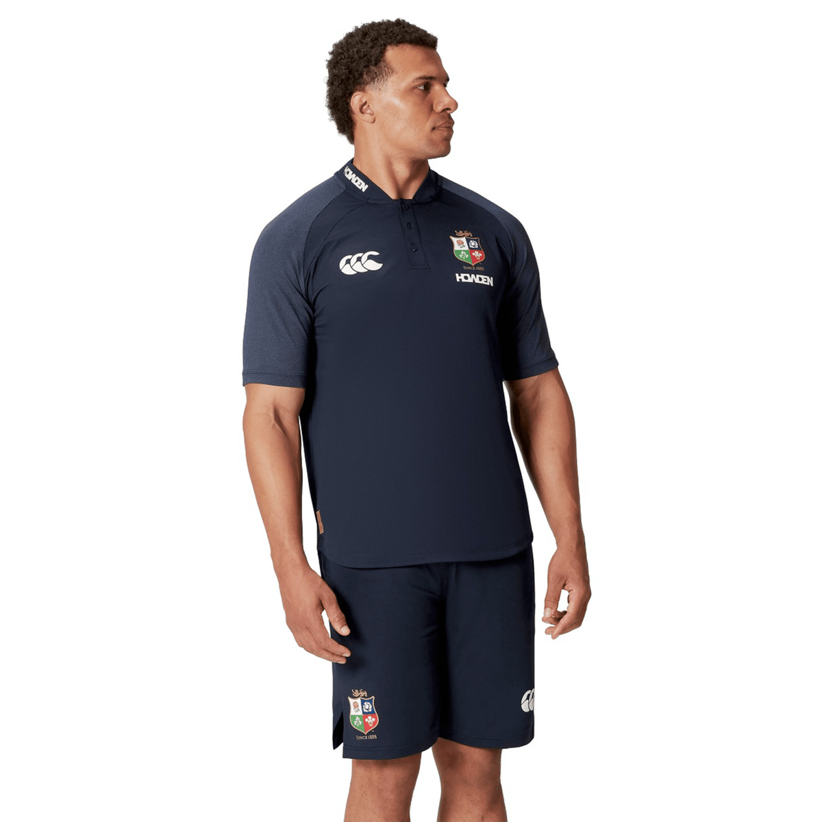 A man wearing the British & Irish Lions 25 Poly Polo, a navy blue sports shirt featuring the Canterbury logo and designed with breathable wicking fabric, stands against a white background.