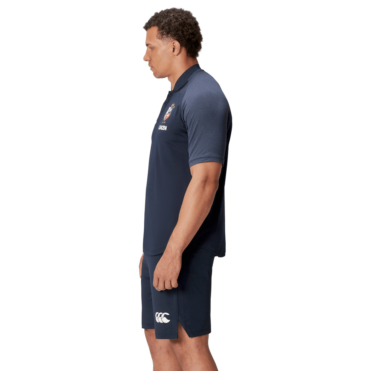 A man wearing the British & Irish Lions 25 Poly Polo by Canterbury in navy, designed with breathable wicking fabric, stands sideways against a white background.
