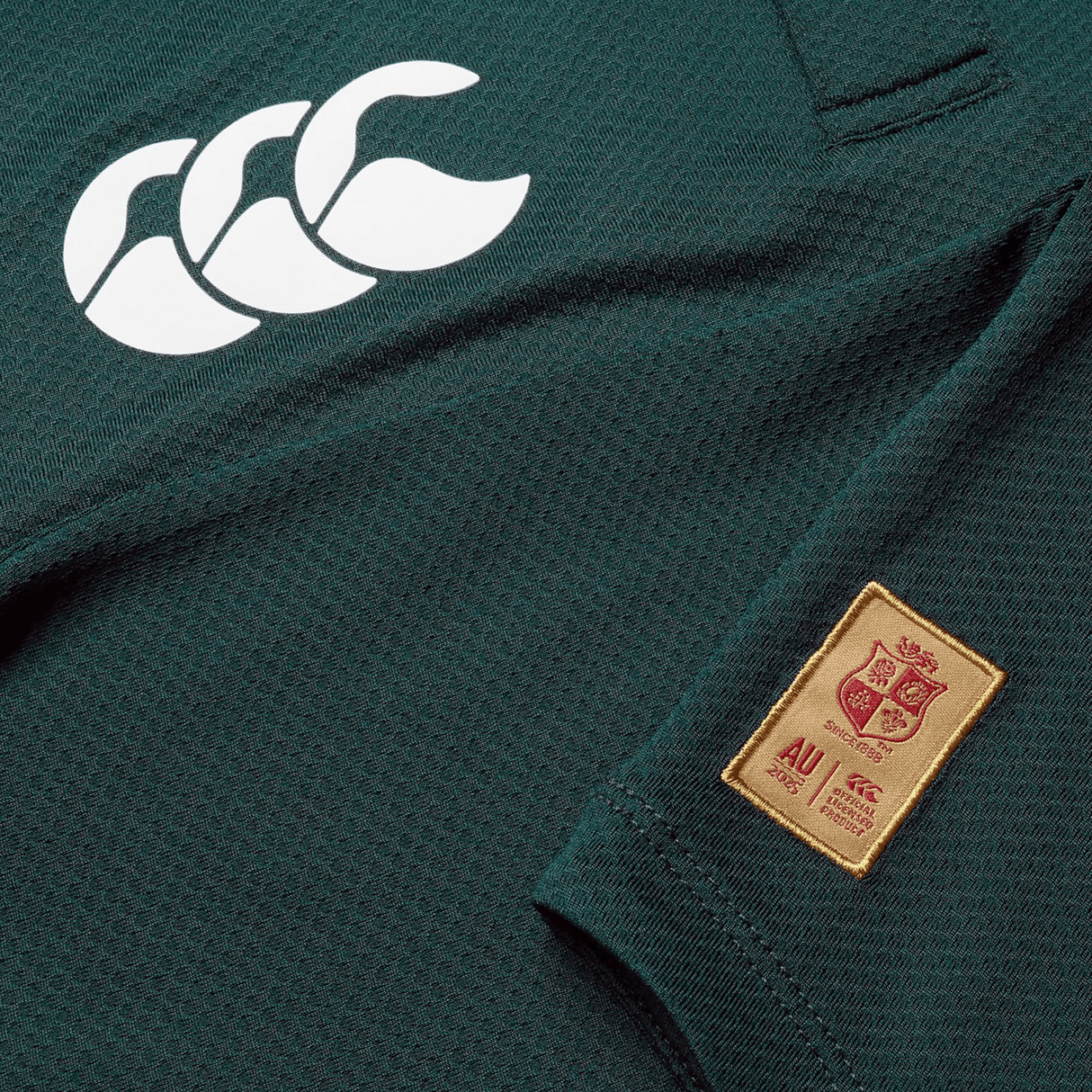 Close-up of a green sports jersey, crafted by Canterbury, featuring a white logo with three overlapping outlines and a red and gold patch displaying a crest and text. This British & Irish Lions 25 Poly Polo is made from quick-drying fabric.