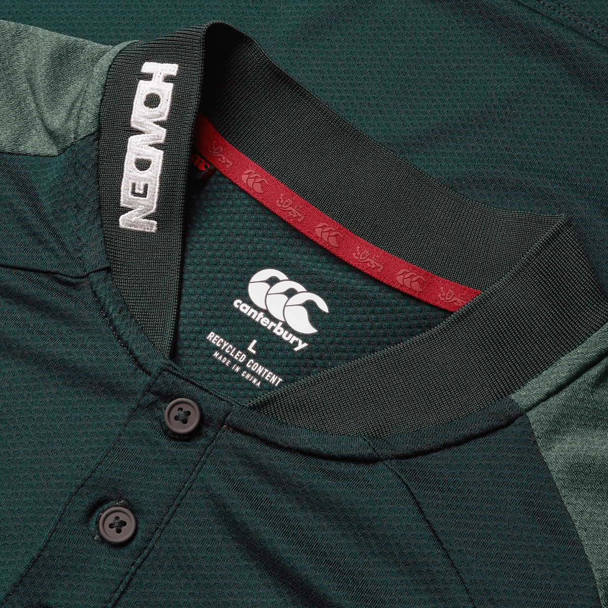 Close-up of a green British & Irish Lions 25 Poly Polo by Canterbury with black buttons, featuring "Howden" on the collar and a "recycled content" label inside. This quick-drying polo is ideal for those who value both performance and sustainability.