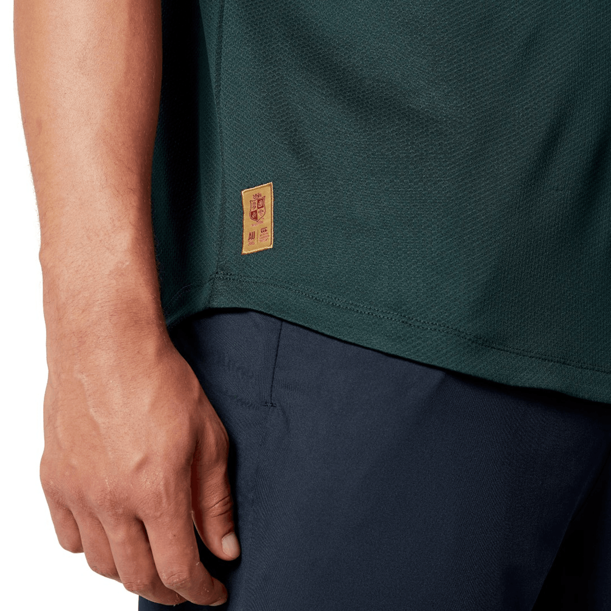 Close-up of a person wearing the British & Irish Lions 25 Poly Polo by Canterbury, featuring a quick-drying dark green fabric and complemented by navy blue pants. A small patch is visible on the shirt as the individual's hand rests by their side.
