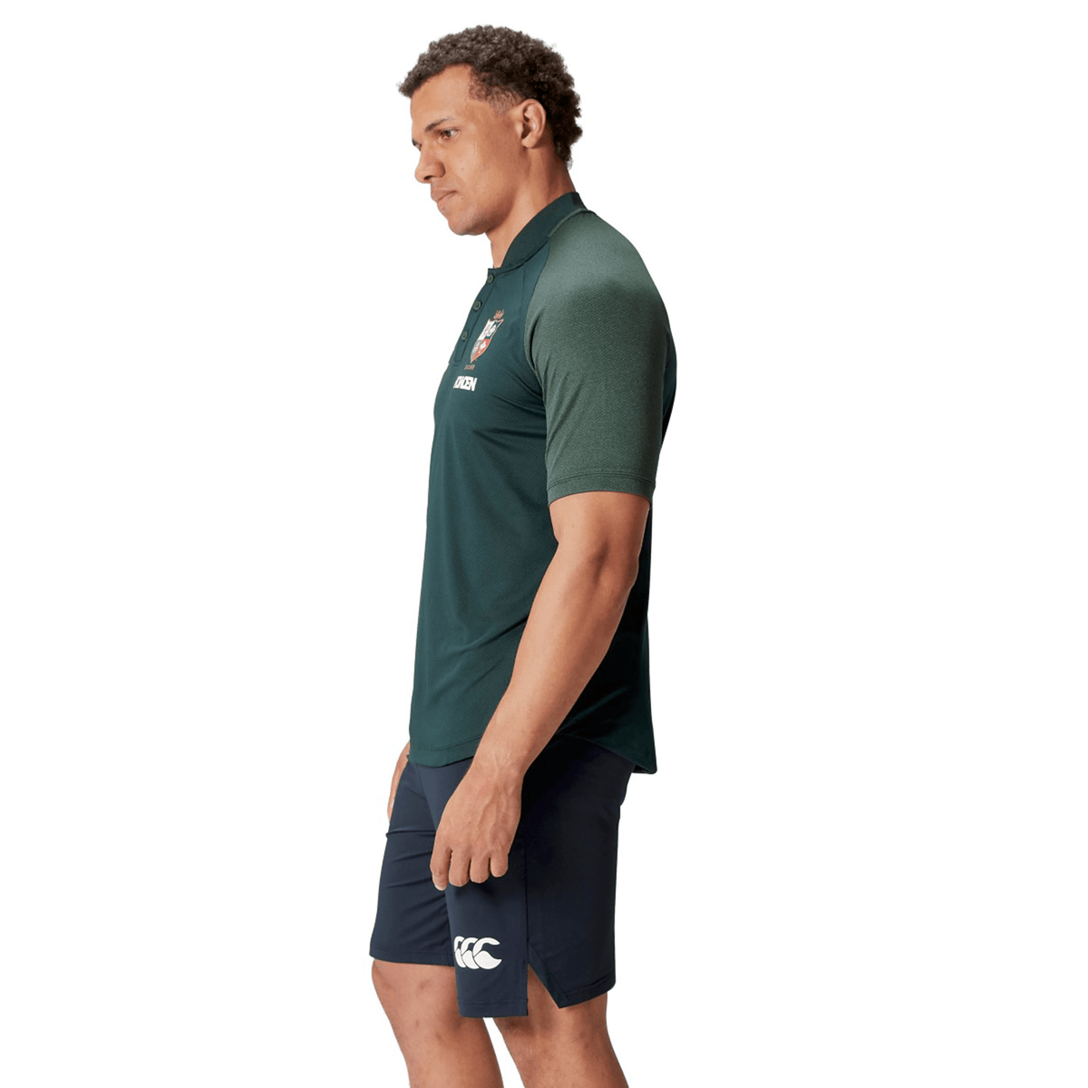 A man wearing the British & Irish Lions 25 Poly Polo by Canterbury, a green collared sports shirt crafted from breathable wicking fabric, and navy shorts stands in a side profile against a white background.